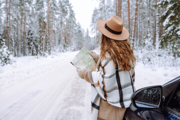 Happy woman in a hat with a map near the car on a snowy winter road in the forest. A traveler is looking for directions using a map on a winter day. Car travel concept.