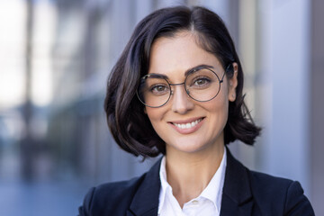 Close-up photo of a young beautiful successful female lawyer in a suit and glasses smiling at the camera and standing outside