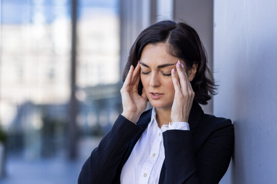 Close-up photo of a young tired businesswoman standing on the street and holding her head, feeling pain and pressure, doing a massage with her hands, closed her eyes