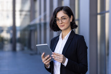 Portrait of a self-confident business woman, executive director, company founder standing in a suit and glasses near the office center, holding a tablet in her hands and smiling at the camera