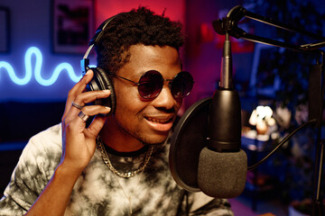 Young male host in headphones talking in microphone in studio illuminated with neon lights while...