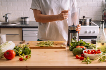Lady in the kitchen throws celery into a blender on a wooden table on which there are fresh...