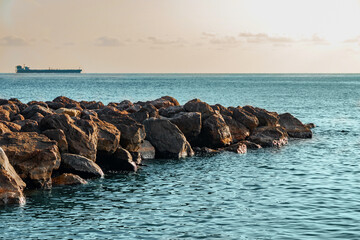 View of the sea breakwater made of stones. Seascape at sunset under a cloudy sky. Waves on the sea and dark clouds in the blue sky. A beach with a breakwater made of large stone boulders. 