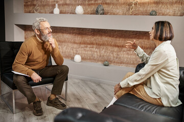 bearded middle aged psychologist in glasses talking to female client during therapy session - obrazy, fototapety, plakaty
