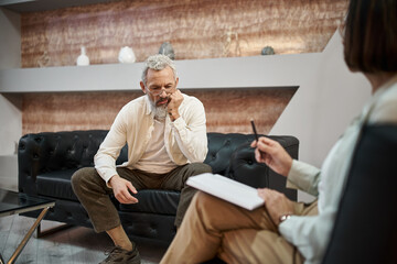 sad bearded middled aged man with tattoo sitting on leather couch near female psychologist