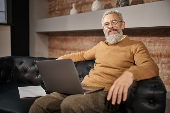 joyful middle aged psychologist with beard sitting on leather couch with laptop, online consultation