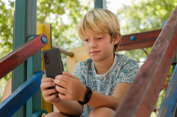 10 years old cute blonde boy using smartphone outdoors. Portrait of concentrated kid sitting in a...