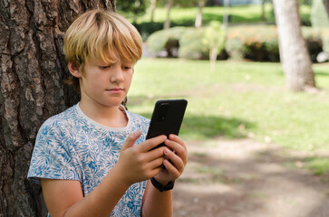 10-year-old handsome boy with blond hair using smartphone outdoors. Portrait of kid leaning against a tree in a park holding mobile phone playing game. Rest, education, technology concept. Copy space.