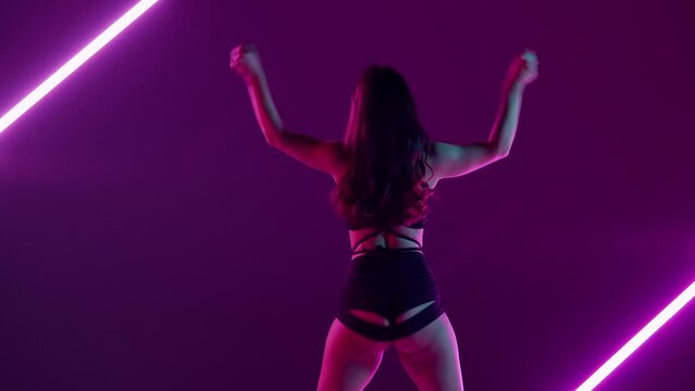 In the frame on a purple, neon background, with bright lights stands a young, slender woman, Demonstrates a dance move in the style of twerk. She is dressed in open clothes, sexy, rhythmic, plastic