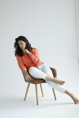 Beautiful woman of 30 years old sitting on chair, white background. Portrait of brunette girl smile. Orange sweater and light jeans. Woman barefeet, thoughtful.