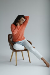 Fototapeta na wymiar Beautiful woman of 30 years old sitting on chair, white background. Portrait of brunette girl smile. Orange sweater and light jeans. Woman barefeet, thoughtful.