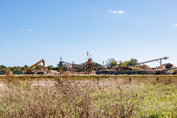 Dutch field in Maasvallei nature reserve, earth, sand and gravel extraction work site with heavy industrial machinery, conveyor belt and crusher in background, sunny day in Meers, Elsloo, Netherlands