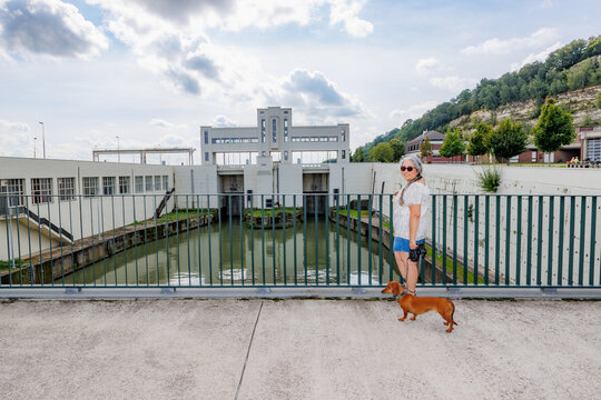 Smiling female tourist together with her dachshund standing in front of Lanaye lock against gray blue sky, Albert canal, reflection in water surface, sunny summer day in Ternaaien, Belgium