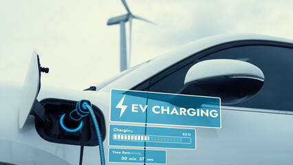 Electric car recharging energy from EV charging station display futuristic smart battery status hologram by EV charger plug cable in wind turbine farm. Alternative clean energy sustainability. Peruse