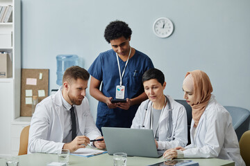 Diverse group of doctors using laptop computer together in clinic with mature woman talking to...