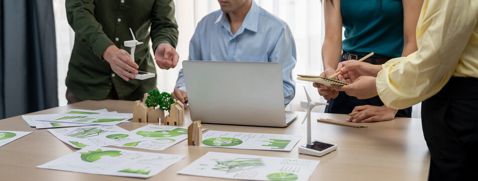 Cropped image of skilled businessman presented eco-friendly house project to general manager at meeting table with wooden block, windmill model and environmental document scatter around. Delineation.