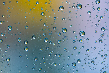 Window pane with many small raindrops and unfocused scenery background in November. Rainy day seen through glass with clear drips, macro close up. Colorful blurred background, yellow-blue gradient 