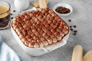 Tradition italian layered dessert tiramisu with mascarpone cream and biscuits in a white baking dish on a gray concrete background.