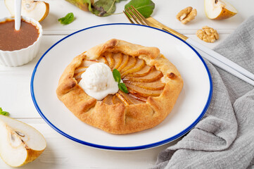 Autumn galette with pear, ice cream, walnuts, caramel sauce and cinnamon on a white wooden background.