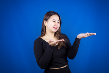 Fototapeta premium An excited young woman presenting and pointing upwards the copy space, isolated on blue background