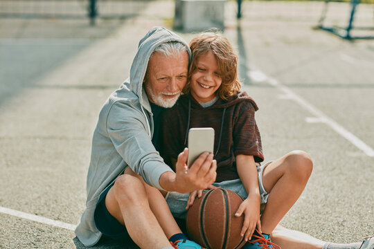 Grandfather and grandson taking a selfie on a smart phone while resting from playing basketball outdoors on a basketball court in the city