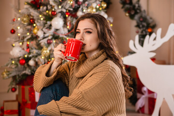 Beautiful young woman with a red mug of coffee in her hands against the background of a Christmas tree.