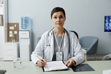 Front view portrait of mature woman as female doctor sitting at workplace in clinic and looking at...