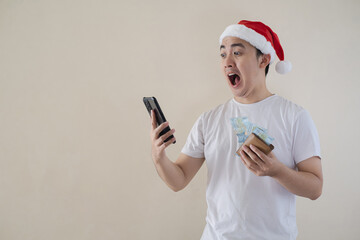 Young Asian Santa man is excited with happy face expression while looking at the smartphone and holding wallet and moneys. Isolated beige background. Christmas concept.