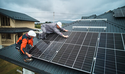 Workers installing photovoltaic solar panels on roof of house. Men engineers in helmets building...