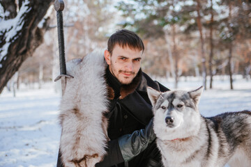 A man in a black cloak with a sword and skin. Warrior in the forest with a wolf in winter.