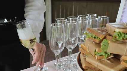 A young woman pours champagne into glasses. Champagne in a glass. Catering with sandwiches. A woman pours champagne into a glass, close-up.