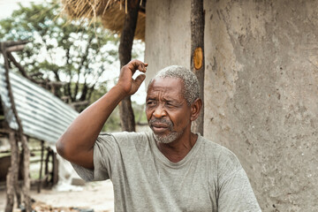 old retired african man scratching his head, some unexpected problem, in front of the house with...