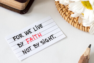 We live by faith, not by sight, handwritten Christian quote on paper with pencil, flowers, and closed holy bible book. Biblical concept of trust in God Jesus Christ, 2 Corinthians 5:7 text.