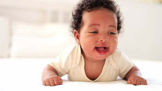 laughing portrait close-up of a small African-American baby girl in a white bodysuit on the bed at home, a funny six-month-old smiling and joyful black newborn baby lying on her tummy