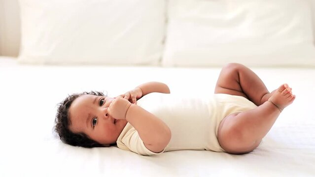 a close-up portrait of a small African-American baby girl in a white bodysuit on a cotton bed at home, a funny six-month-old smiling joyful black newborn baby lies on the back