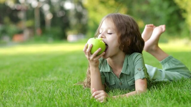 a baby girl eats a green apple on a green lawn in summer and smiles lying on the grass, a cheerful child with an apple, children's holidays, close-up, healthy teeth