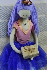 A beautiful rag doll with purple hair, a pink blouse and a blue skirt holds in her hands a cardboard box with a gift parcel tied with a rope. 