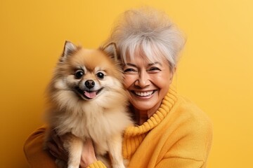 Portrait of smiling Caucasian senior woman in yellow sweater posing with cute Pomeranian spitz puppy. Happy lady hugs her beloved pet. Love between human and dog. Yellow studio background, copy space.