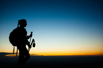 Fototapeta na wymiar Silhouette of a hiker girl on a rock pedestal with hands up. Beautiful orange sunset on blue sky.