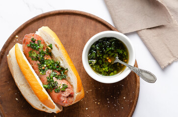 A choripan with chimichurri sauce on a wooden board and a brown napkin on white marble background.
