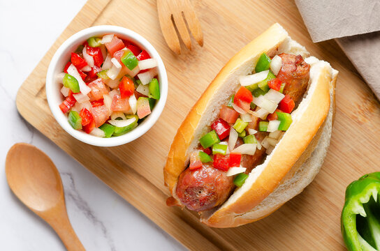 A choripan with Argentinian criolla sauce on a wooden board, a green pepper, wooden fork and spoon, and a brown napkin on white marble background.
