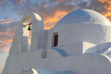 Sunset starts on the island of Mykonos with a white-washed church with a lone bell aglow in the foreground. 