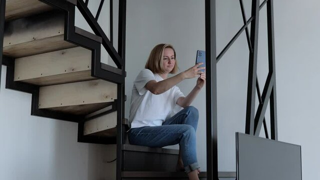 A woman takes a photo with a smartphone. A young woman is sitting on the stairs in an apartment, with a smartphone in her hands. Woman at home alone, sitting on a ladder