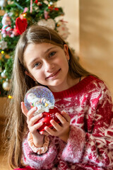 Naklejka premium Radiant 8-Year-Old Girl, Joyful with a Snow Globe by the Christmas Tree at Home