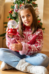 Cheerful 8-Year-Old Girl Grins by the Christmas Tree and Snow Globe