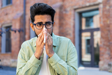 A man sneezes while sitting on a bench outside an office building, a young businessman has a runny nose and allergies, sick - obrazy, fototapety, plakaty