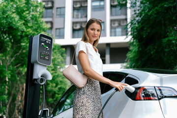 Young woman travel with EV electric car charging in green sustainable city outdoor garden in...