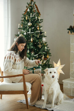 Woman in cozy sweater opening christmas gift with cute white dog on background of stylish christmas tree and fireplace. Winter holidays. Owner with pet in festive room. Merry Christmas!