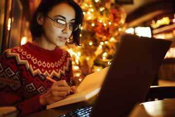 Young busy woman in red xmas sweater working in cozy cafe before Christmas Eve. She is seated in front of a laptop, making notes in a notebook