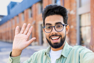 Phone web camera view, young man uses app on smartphone for remote video call communication, Indian man smiles and looks at camera, waves hand in greeting